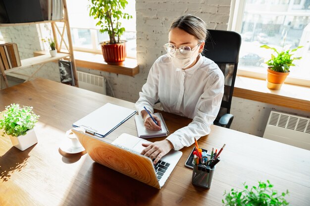 Reunión. Mujer que trabaja sola en la oficina durante la cuarentena por coronavirus o COVID-19, con mascarilla. Joven empresaria, gerente haciendo tareas con teléfono inteligente, computadora portátil, tableta tiene conferencia en línea.