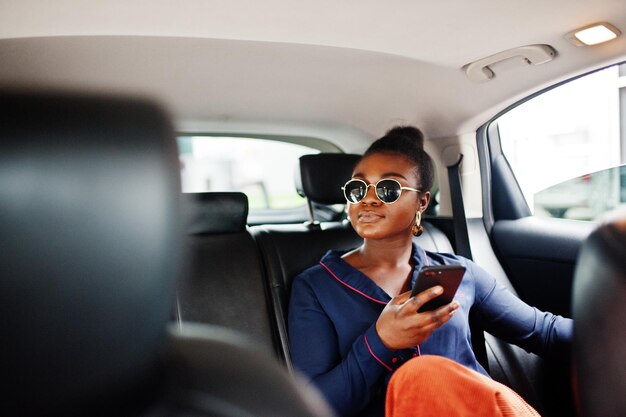 Una rica mujer africana de negocios con gafas de sol se sienta en un auto todoterreno con asientos de cuero negro Teléfono móvil a mano