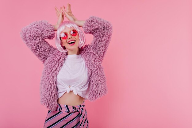 Riendo agradable chica con tatuaje posando en periwig sobre fondo rosa Foto de estudio de éxtasis modelo femenino blanco viste elegante chaqueta de piel y gafas