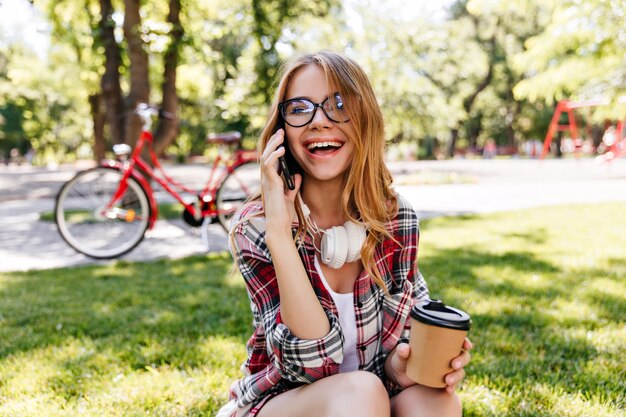 Riendo alegre niña hablando por teléfono en el parque. Debonair mujer europea con gafas llamando amigo en día de verano.