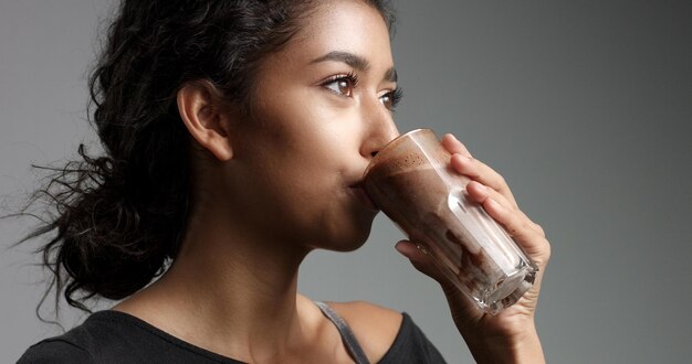 Riendo atractiva chica del Medio Oriente en la parte superior negra relajada bebiendo café mocca chocolate caliente de un vaso alto aislado en gris