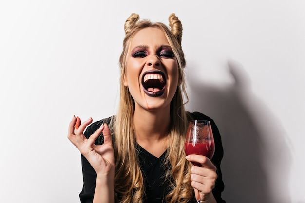 Riendo bruja malvada posando con poción. Vampiro rubio alegre bebiendo sangre en la fiesta de halloween.