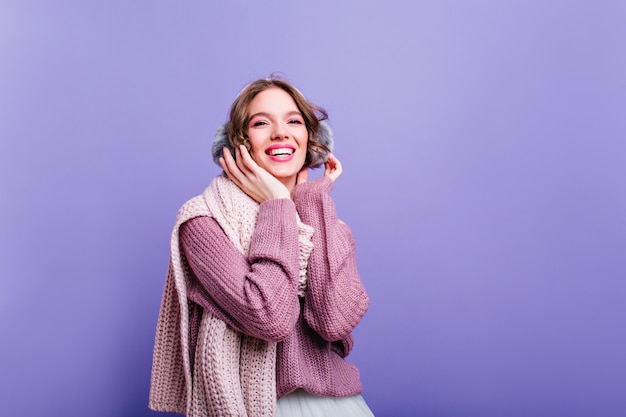 Riendo a la chica de moda en auriculares de invierno posando Foto interior de mujer feliz inspirada en elegantes accesorios sonriendo en la pared púrpura.