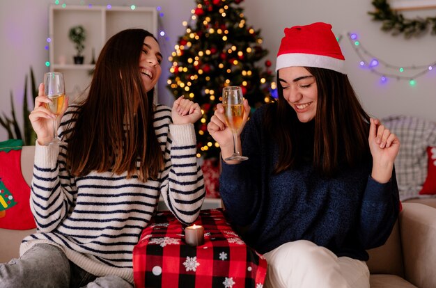 Riendo chicas guapas sostienen copas de champán sentadas en sillones y disfrutan de la Navidad en casa