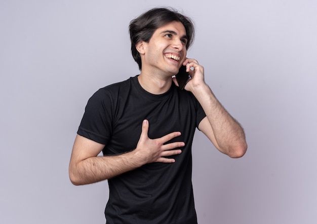 Riendo chico guapo joven con camiseta negra habla por teléfono poniendo la mano en el pecho aislado en la pared blanca