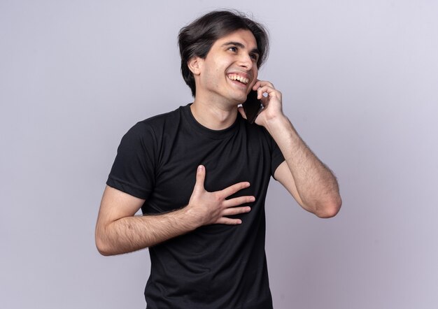 Riendo chico guapo joven con camiseta negra habla por teléfono poniendo la mano en el pecho aislado en la pared blanca