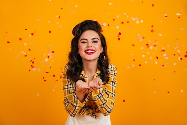 Riendo a dama europea posando con confeti. Chica pinup divertida de pie sobre fondo amarillo con una sonrisa sincera.