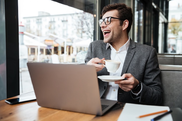 Riendo empresario en anteojos sentado junto a la mesa en la cafetería con computadora portátil y una taza de café mientras mira a la ventana