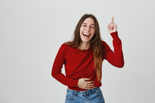 Riendo feliz mujer atractiva, tocando el vientre y apuntando con el dedo hacia arriba