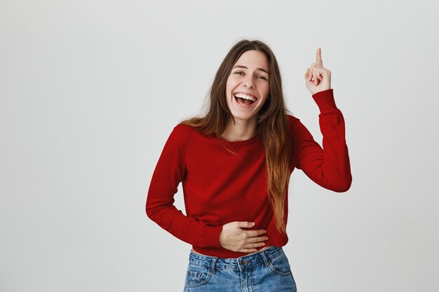 Riendo feliz mujer atractiva, tocando el vientre y apuntando con el dedo hacia arriba