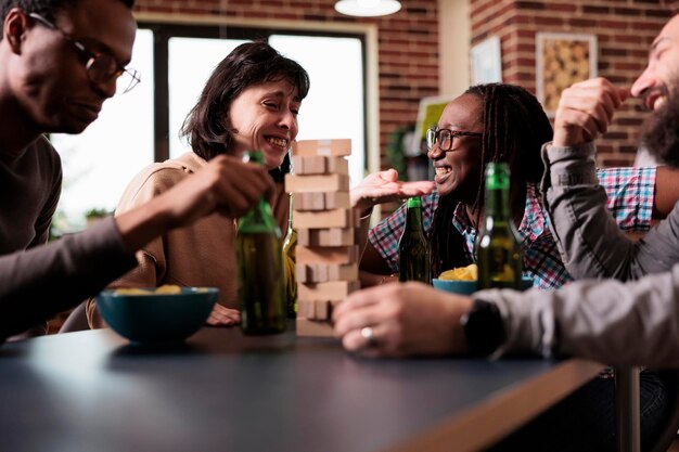 Riendo a un grupo diverso de amigos sentados a la mesa en la sala de estar mientras juegan juegos de sociedad. Personas multiétnicas casuales sentadas en casa mientras disfrutan juntos de divertidas actividades de ocio.