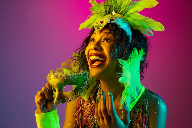 Riendo. Hermosa mujer joven en carnaval, elegante disfraz de mascarada con plumas bailando sobre fondo degradado en neón. Concepto de celebración navideña, tiempo festivo, baile, fiesta, diversión.