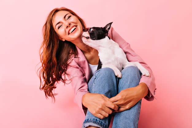 Riendo jocund mujer sentada en el suelo con perro de rodillas. Retrato de interior de dama agradable posando con cachorro en pastel.