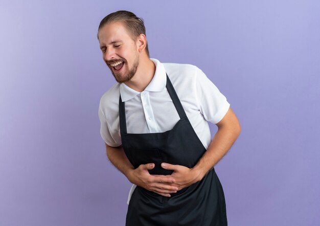 Riendo joven barbero guapo vistiendo uniforme sosteniendo su vientre con los ojos cerrados aislado sobre fondo púrpura espacio de copia