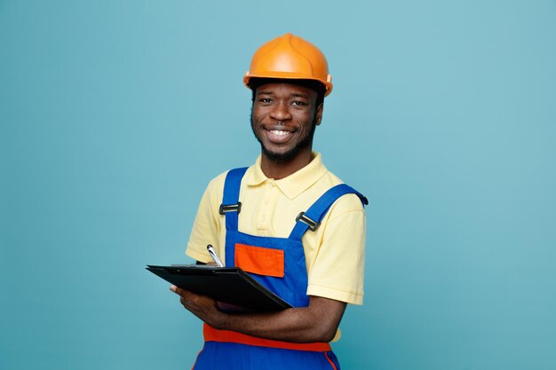 Riendo joven constructor afroamericano en uniforme sosteniendo portapapeles aislado sobre fondo azul.