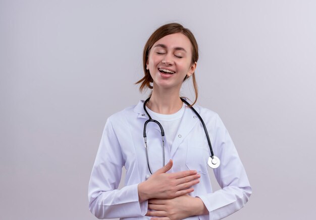 Riendo joven doctora vistiendo bata médica y estetoscopio y poniendo las manos sobre el vientre con los ojos cerrados con espacio de copia