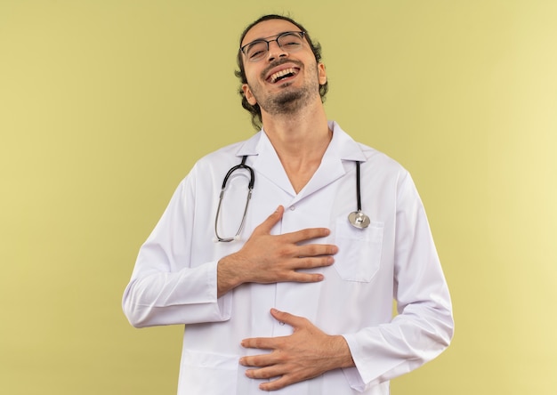 Riendo joven médico con gafas ópticas vistiendo bata blanca con estetoscopio poniendo las manos en el pecho y el estómago