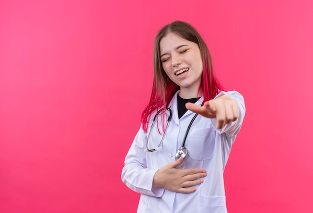 Riendo joven médico mujer vistiendo estetoscopio bata médica que le muestra gesto en rosa pared aislada