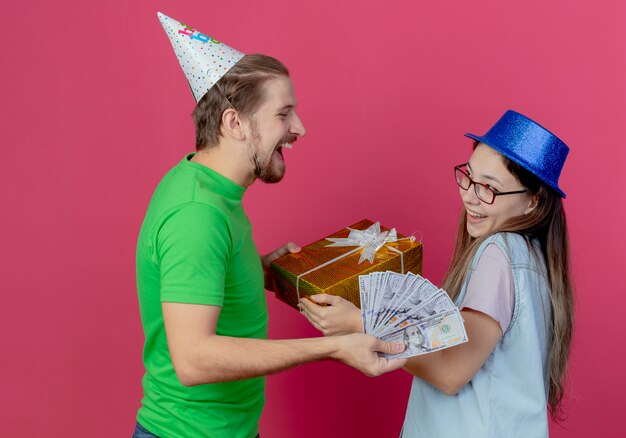 Riendo joven vistiendo gorro de fiesta ofrece dinero a una joven complacida con gorro de fiesta azul para llevar caja de regalo aislada en la pared rosa