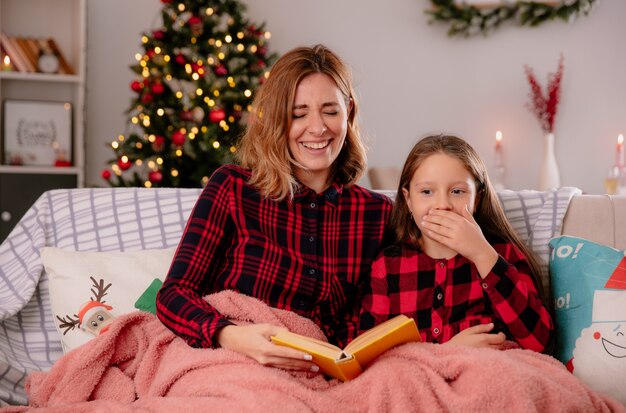 Riendo, madre e hija, libro de lectura cubierto con una manta sentada en el sofá y disfrutando de la Navidad en casa