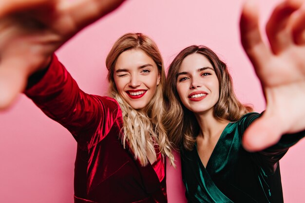 Riendo a mujer caucásica con lápiz labial rojo haciendo selfie en fiesta. Atractivas amigas relajándose juntas.