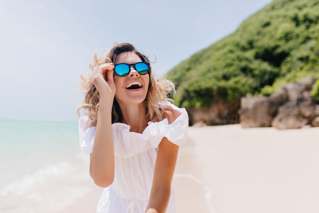 Riendo mujer espectacular con gafas de sol disfrutando de vacaciones en la isla tropical. Foto al aire libre de mujer adorable en vestido blanco sonriendo en la naturaleza.