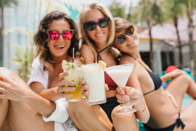 Riendo a mujer morena con gafas de sol rosas celebrando algo con amigos durante el descanso de verano. Hermosas damas bronceadas bebiendo cócteles y disfrutando de las vacaciones.