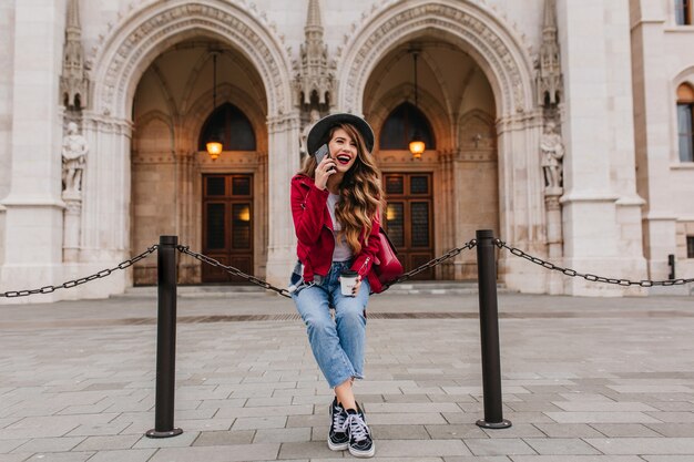 Riendo a mujer de pelo largo en jeans y chaqueta roja sentada frente al antiguo museo hermoso