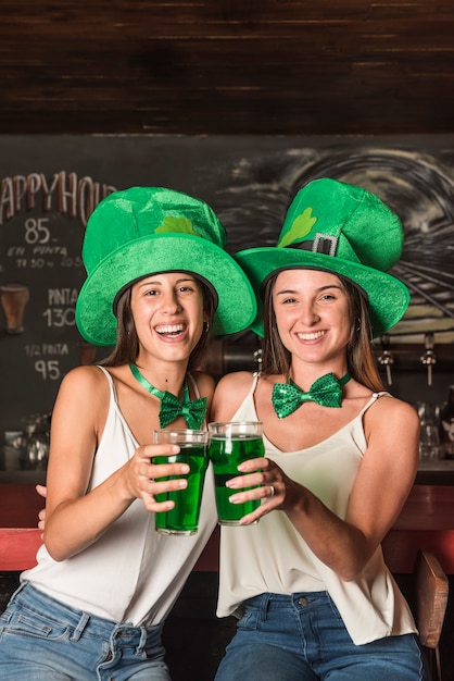 Foto gratuita riendo a mujeres jóvenes con sombreros de san patricio abrazando y sosteniendo vasos de bebida en la barra del bar