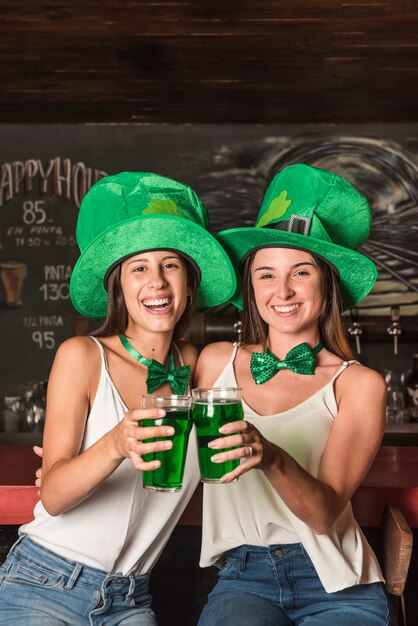 Riendo a mujeres jóvenes con sombreros de San Patricio abrazando y sosteniendo vasos de bebida en la barra del bar