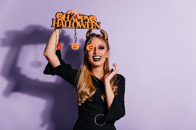 Riendo a la niña blanca en vestido de halloween posando en la pared púrpura. Preciosa modelo femenina en traje de fiesta expresando emociones positivas.
