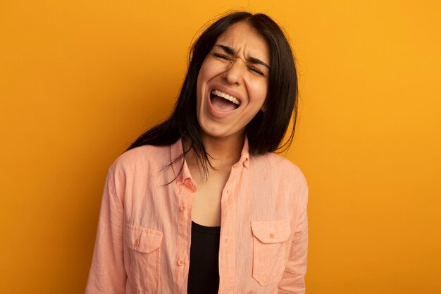 Riendo con los ojos cerrados hermosa joven vestida con camiseta rosa aislada en la pared amarilla