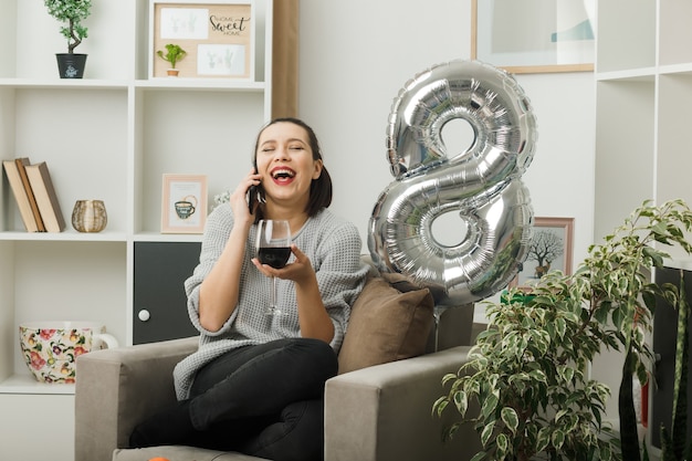 Riendo con los ojos cerrados hermosa mujer en el día de la mujer feliz sosteniendo una copa de vino habla sobre el vino sentado en un sillón en la sala de estar