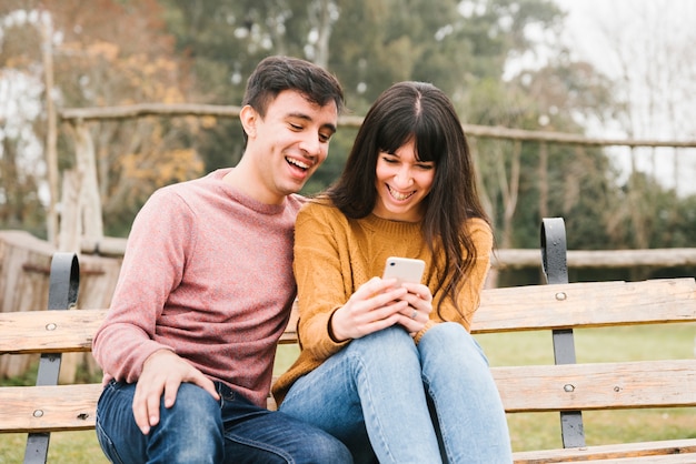 Riendo pareja sentada en el banco y mirando móvil