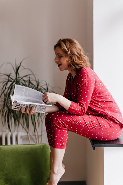 Riendo la revista de lectura de mujer descalza en la mañana. Filmación en interiores de una mujer bonita en pijama sentada en el alféizar de la ventana.