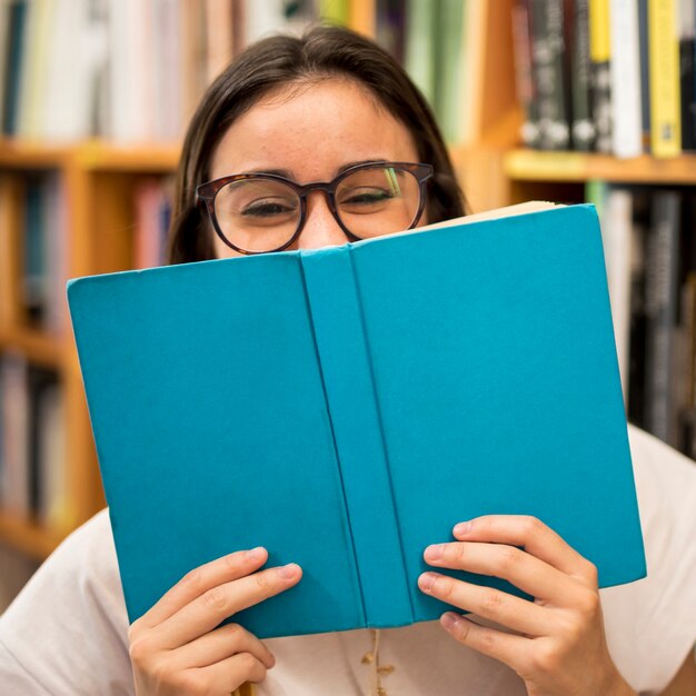 Risa colegiala adolescente escondiendo la cara detrás del libro