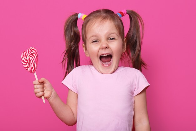 Risa gritando linda chica abre la boca ampliamente, mostrando los dientes, sostiene en una mano corazón sabroso paleta brillante. El niño emocional positivo pasa su tiempo libre con placer. Copie espacio para anuncio.