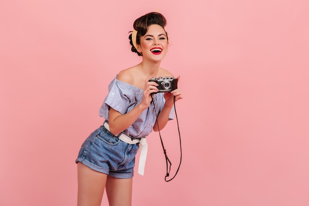 Risa a mujer joven en pantalones cortos de mezclilla tomando fotografías. Foto de estudio de chica pinup con cámara aislada sobre fondo rosa.