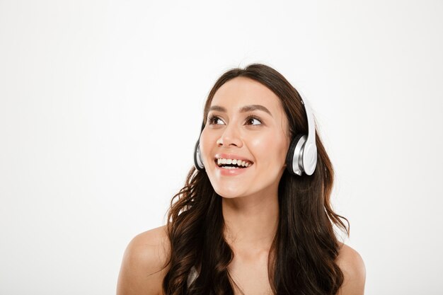 Risa mujer morena en auriculares escuchando música y mirando a otro lado sobre gris