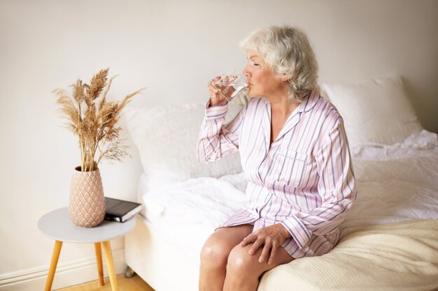 Rituales matutinos, ocio, descanso y concepto de hora de dormir. Atractiva pensionista con canas sentada en la cama en un interior acogedor, sosteniendo una taza, bebiendo agua para que su sistema digestivo funcione