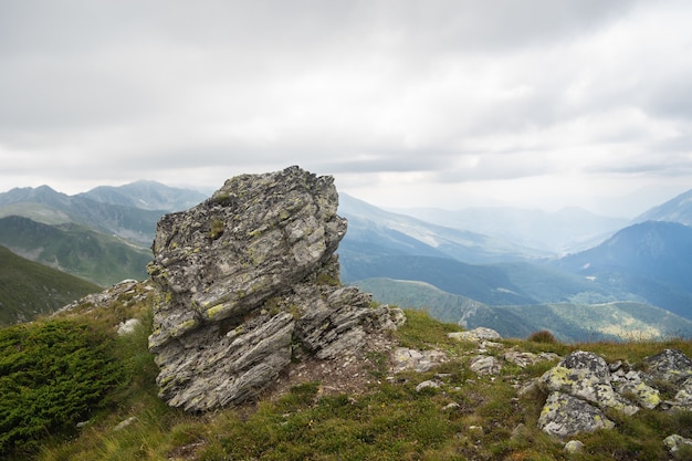 Roca en una colina cubierta de vegetación con montañas rocosas bajo un cielo nublado