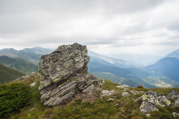 Roca en una colina cubierta de vegetación con montañas rocosas bajo un cielo nublado