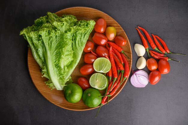 Rodaja de limón, chalotes, ajo, tomates, lechuga y pimientos en un plato de madera sobre un piso de cemento negro.