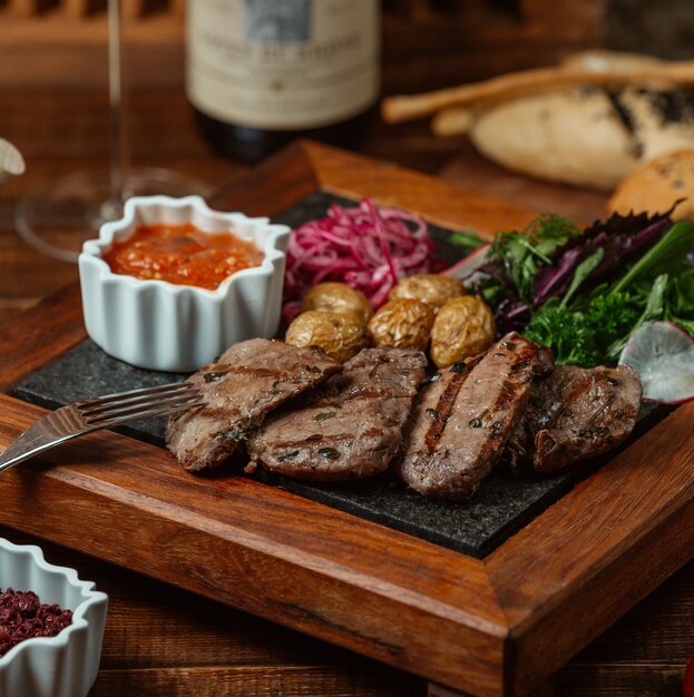 Rodajas de carne marinada a la parrilla servidas con papas baby, cebolla, ensalada de berenjena y hierbas