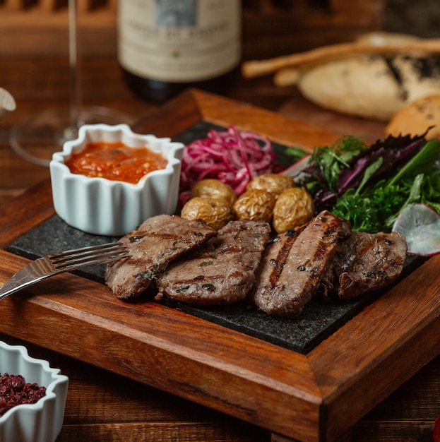 Rodajas de carne marinada a la parrilla servidas con papas baby, cebolla, ensalada de berenjena y hierbas