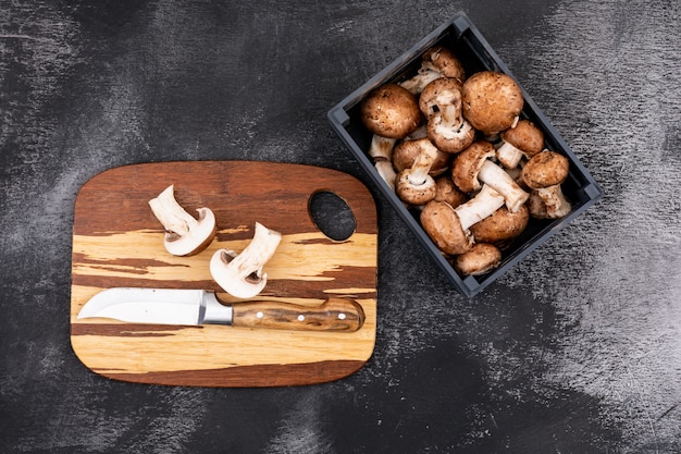 Rodajas de champiñones con un cuchillo en la tabla de cortar de madera cerca de la caja de madera con champiñones frescos