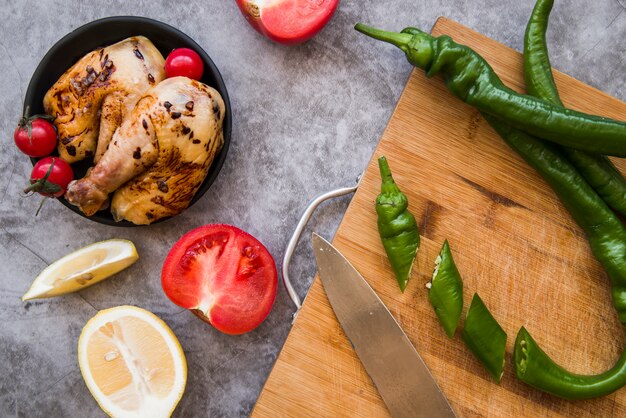 Rodajas de chiles verdes y un cuchillo en una tabla de cortar de madera con pollo a la parrilla; tomate; limón sobre fondo de hormigón