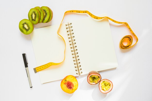 Rodajas de kiwi; durazno y maracuyá con cinta métrica; Pluma y cuaderno espiral sobre fondo blanco