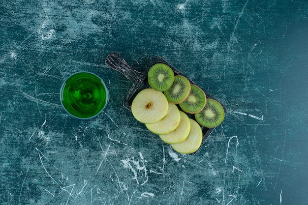 Rodajas de kiwi y manzana en el tablero junto al jugo de estragón, en la mesa azul.