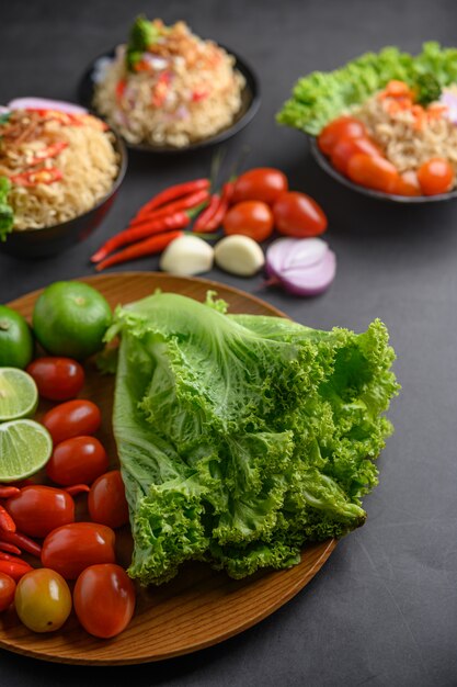 Rodajas de limón, chalotes, ajo, tomates, lechuga y pimientos en un plato de madera sobre un piso de cemento negro.